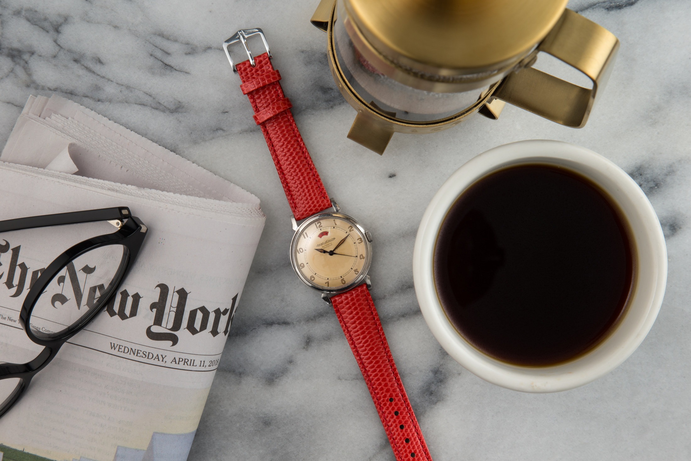 Flat lay photography for 15 hours Vintage watches. Scene: vintage watch with red band on marble counter top surrounded by folded New York Times newspaper, cup of coffee and coffee press.