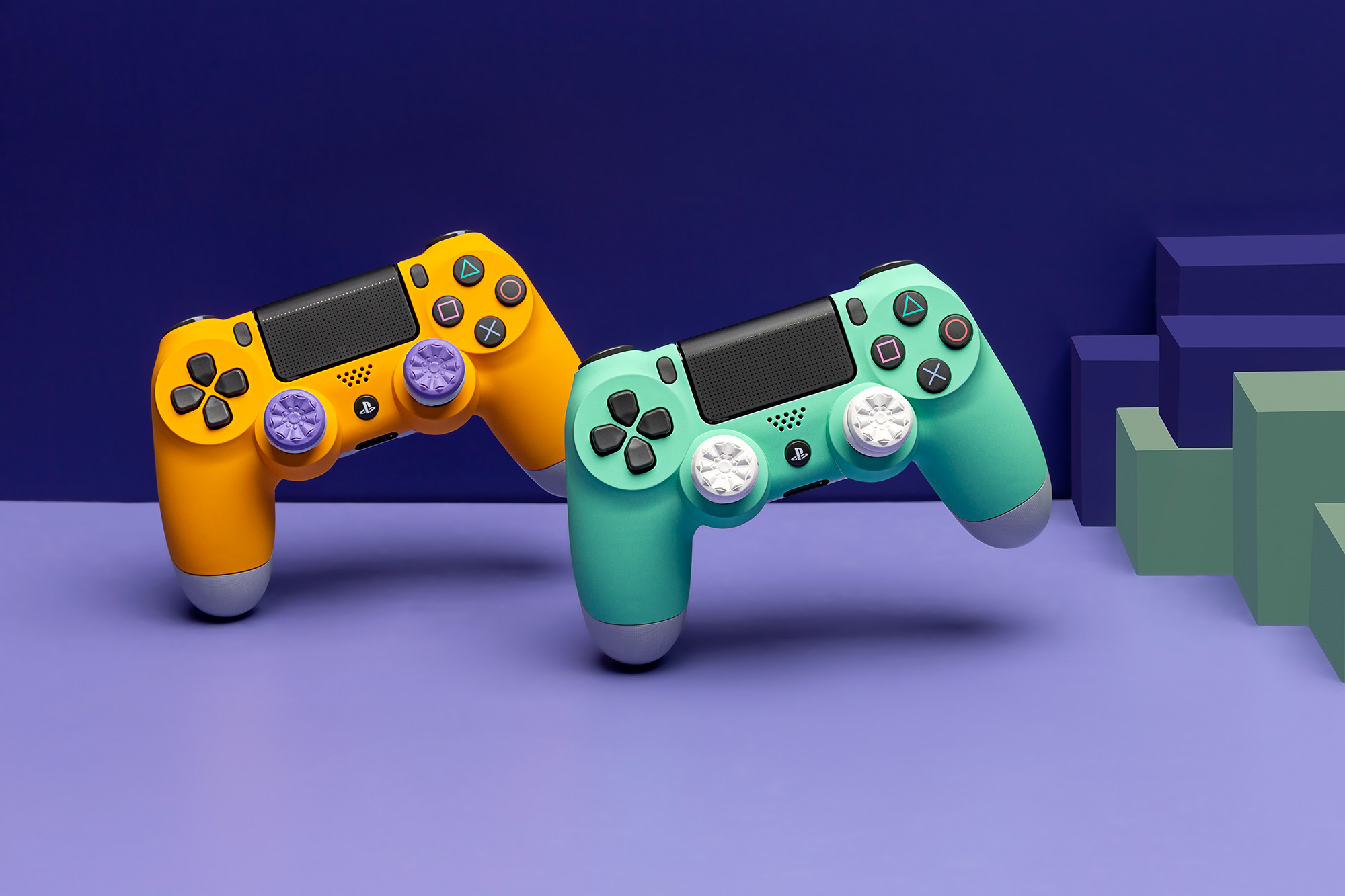 Styled product photography of orange playstation controller with purple thumbsticks next to a mint colored playstation controller with white thumbsticks