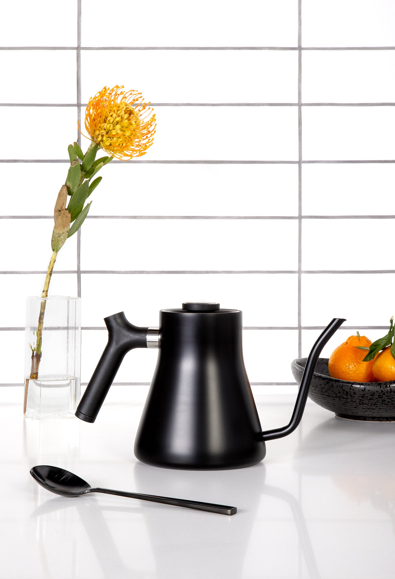 Black teapot on white countertop and white tiled back splash, tea pot is surrounded by a black spoon, black bowl filled with oranges and a single flower in glass vase.